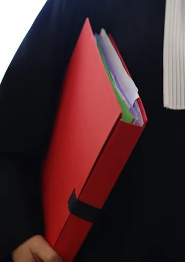 Une avocate ou un avocat en toge noire et rabat blanc, le visage hors cadre, tient un dossier rouge.